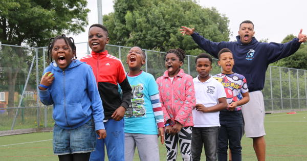 a group of children in a line shouting and smiling on a 5-a-side football pitch