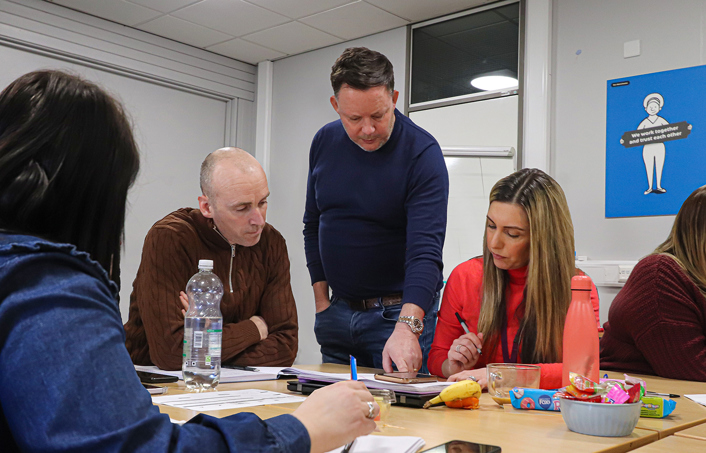 man hosting a seminal standing over people looking at a sheet of paper on a desk